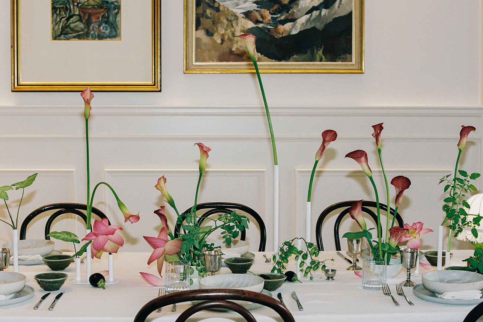 Dining table set with minimal floral arrangements of calla lilies, lotus flower, fruits and cutlery, framed artwork on the wall.