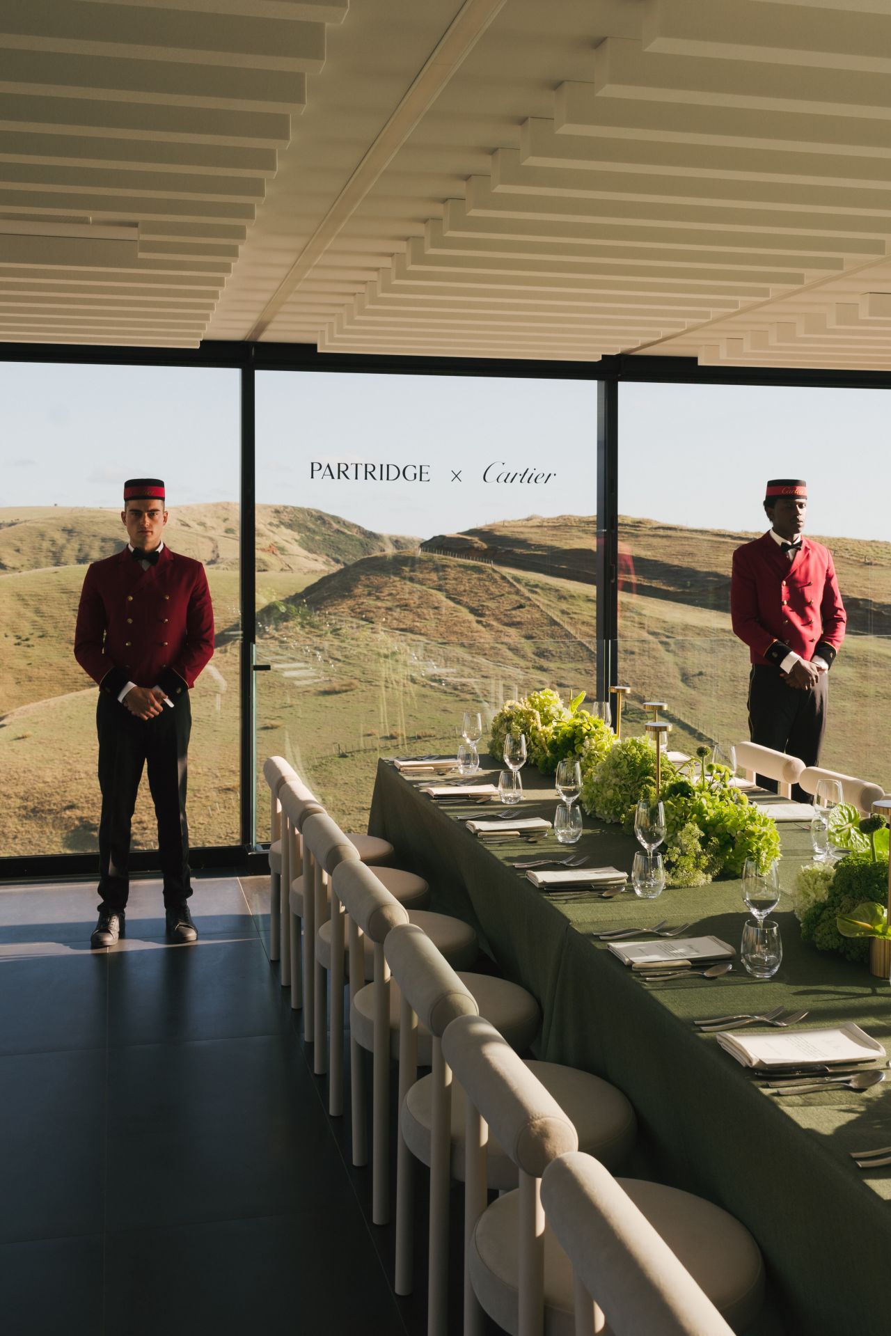 Two people in red uniforms standing in a modern dining area with large windows overlooking a scenic landscape. A table setting with green table cloths and large green floral arrangements running down the table.
