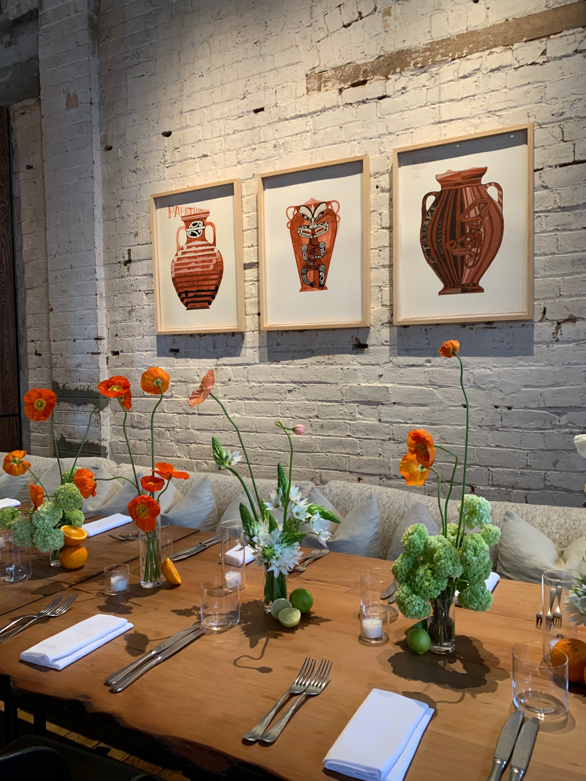 Dining table set with floral arrangements and cutlery against a brick wall with framed vase art. Flowers include poppies, snowball and Ornithogalum