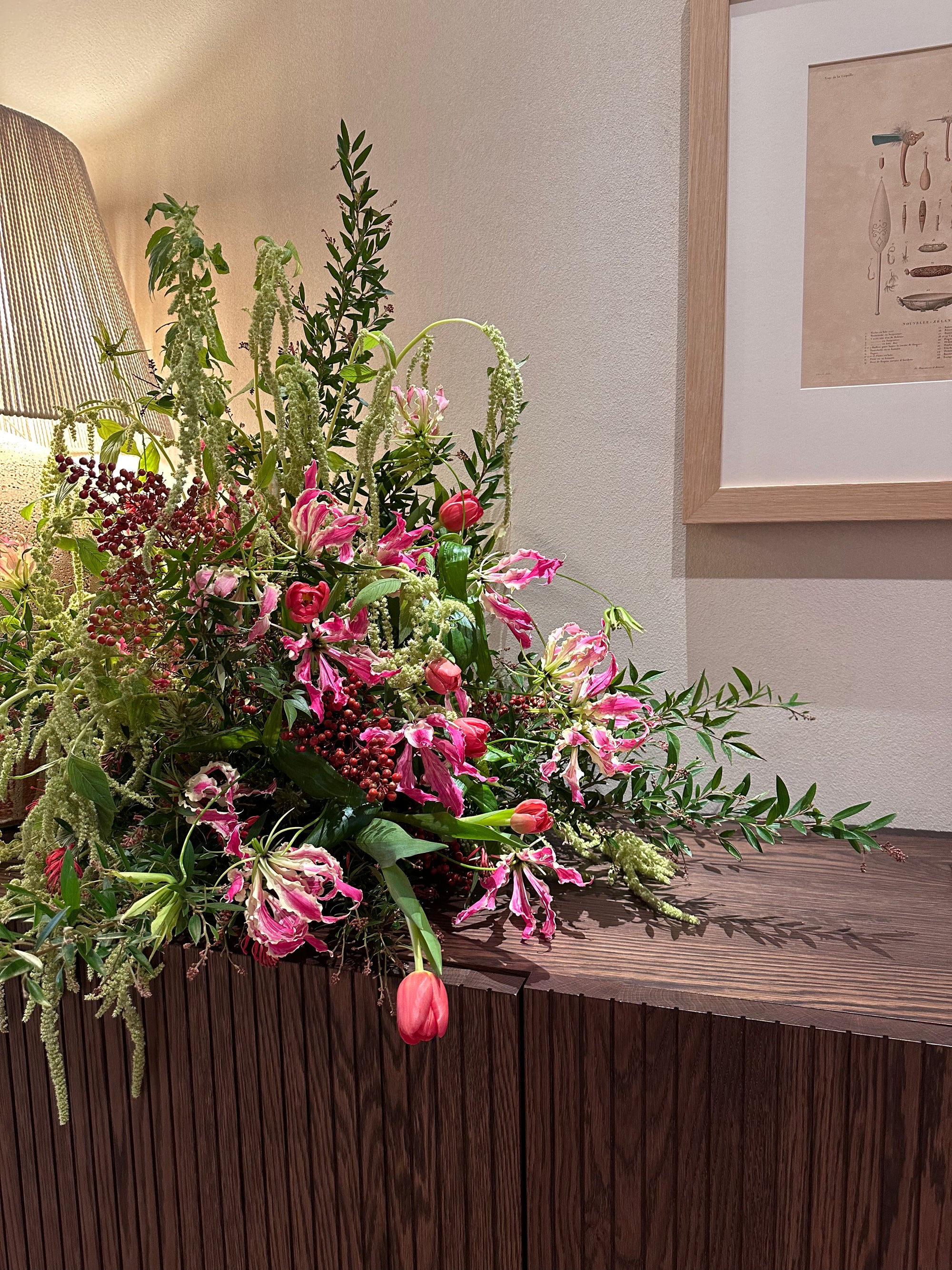 Large floral arrangement on a wooden surface with a lamp and framed picture in the background. Featuring Gloriosa Lilies and Tulips.