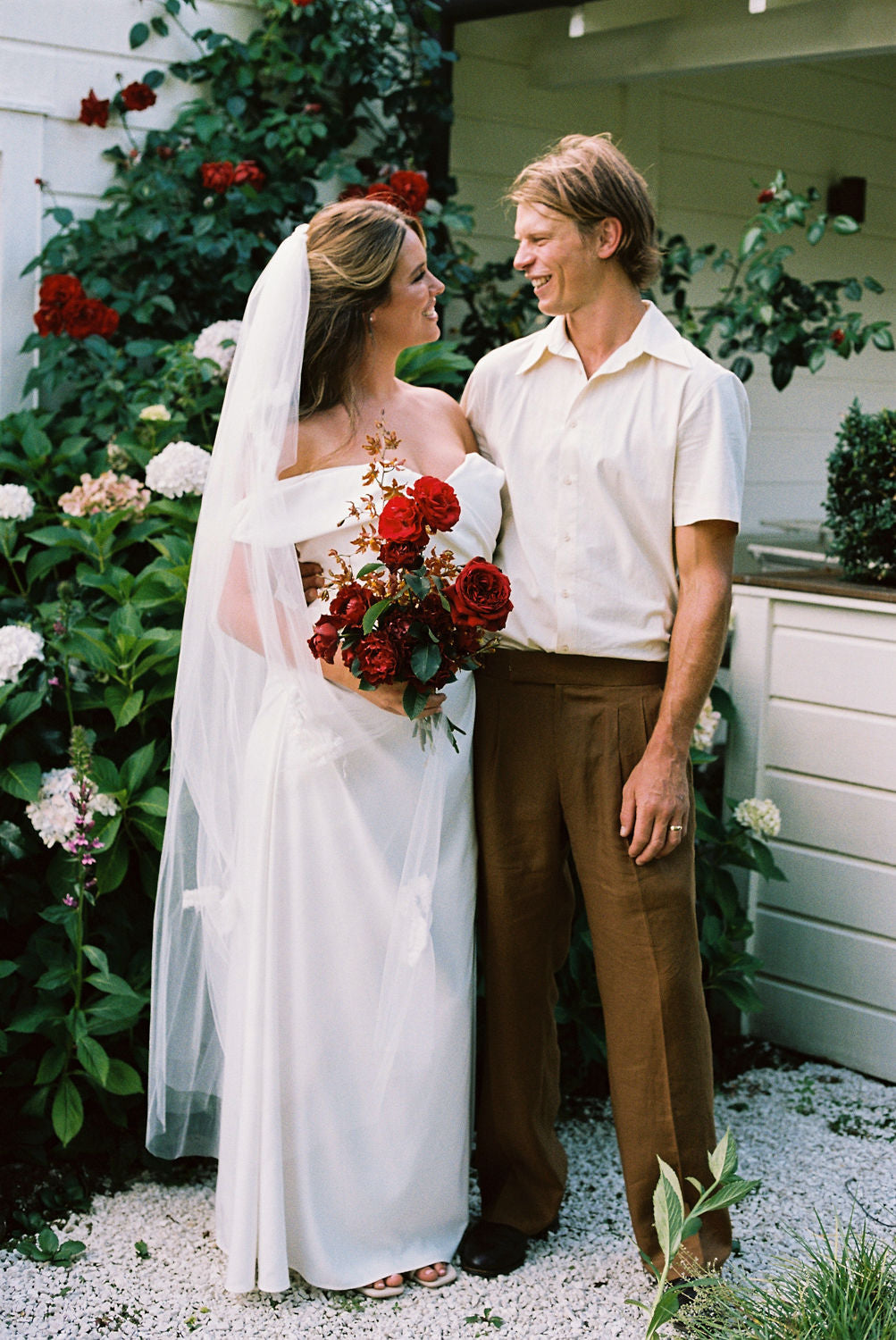 Man and woman standing together on their wedding day outdoors with a white building and greenery in the background while woman is holding a bridal bouquet