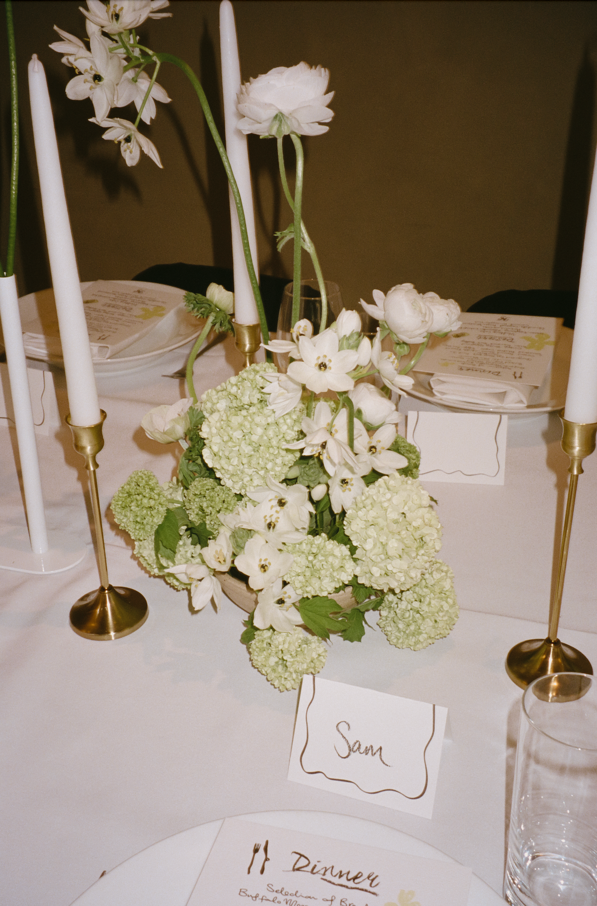 Decorative table setting with white flowers, candles, and place cards on a white tablecloth.